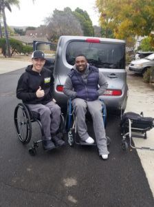 two men in wheelchairs post in front of a vehicle for share-a-chair program