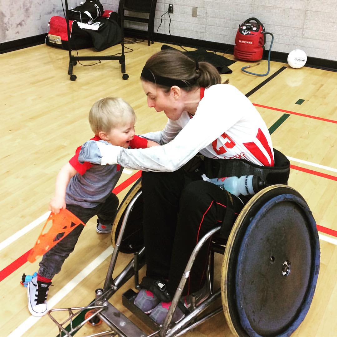Kristen sitting in a rugby wheelchair, playing with and smiling at a child